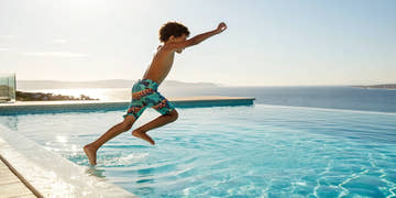 Happy young boy jumping into a pool wearing colorful junior boys swimwear.