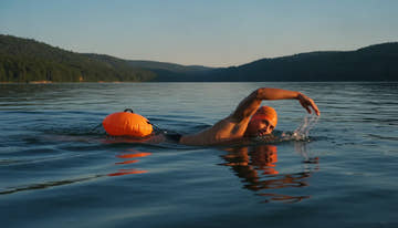Open water swimmer wearing a bright orange swim cap and towing a safety buoy in a lake on a sunny day