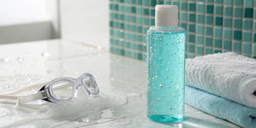 A bottle of TRISWIM shampoo sits poolside with swim goggles and a towel.